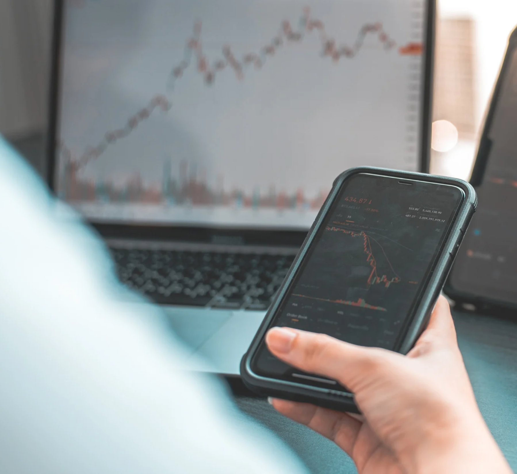 Eine Hand hält ein Smartphone mit Kursverlauf, im Hintergrund ein Laptop mit Chart